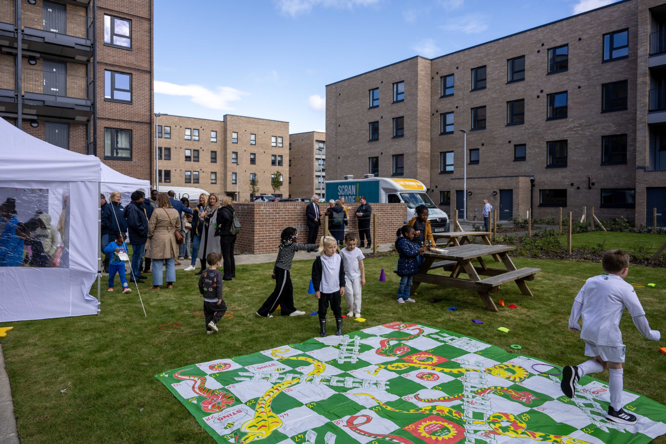Photo of a crowd gathered at a street party with giant snakes and ladders on the grass.