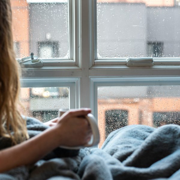 Photo of a person sitting by a window wrapped up in a blanket holding a hot drink in a mug.