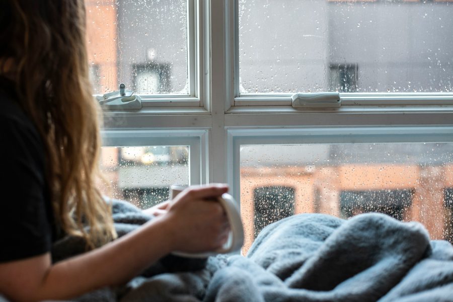 Photo of a person sitting by a window wrapped up in a blanket holding a hot drink in a mug.