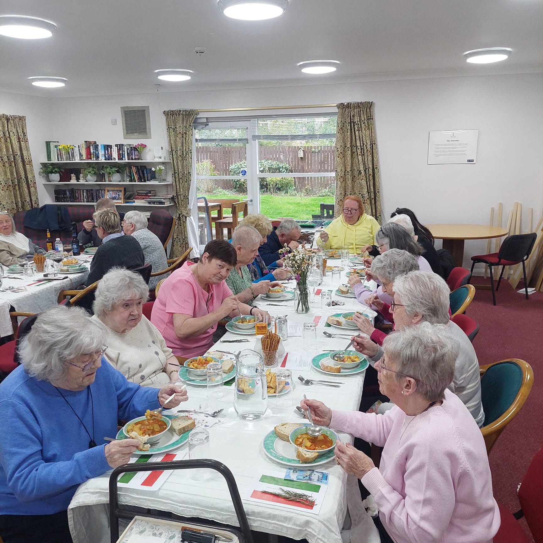 Photo of sheltered housing tenants sitting around a long dining table enjoying a meal. There are Hungarian flags on the table.