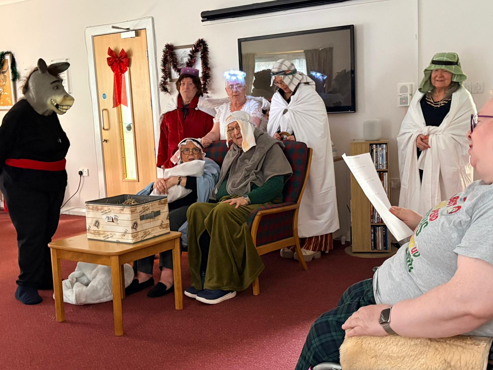 Photo of sheltered housing tenants doing a nativity scene. One tenant wears a donkey head and is accompanied by nativity characters including shepherds, wise men, an angel, Mary, Joseph and baby Jesus. 