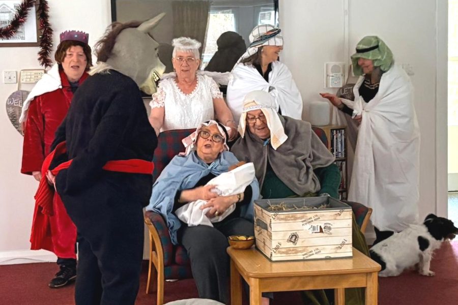 Photo of sheltered housing tenants doing a nativity scene. One tenant wears a donkey head and is accompanied by nativity characters including shepherds, wise men, an angel, Mary, Joseph and baby Jesus. There is also a small black and white dog joining in the scene.